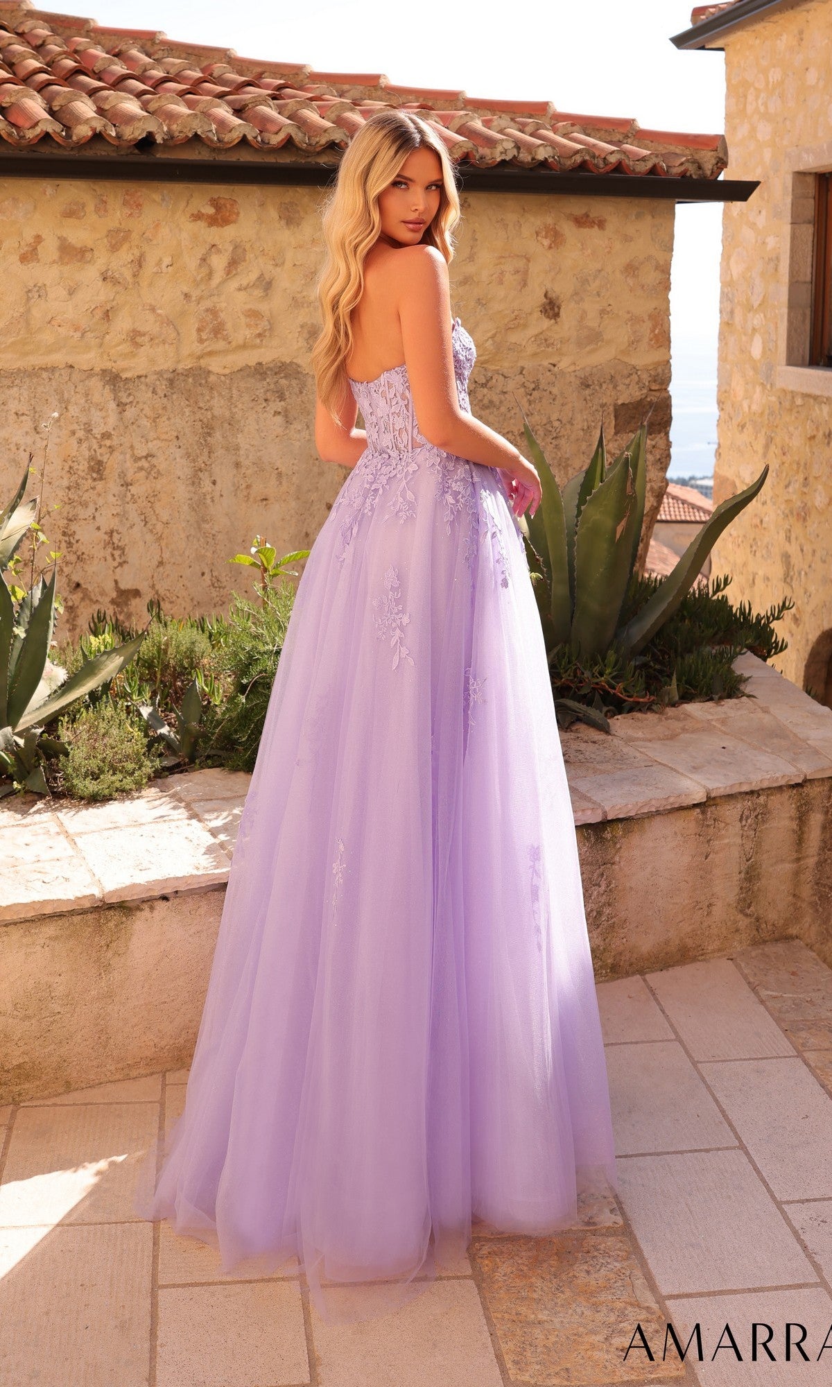 A woman with long blonde hair stands outdoors in front of a stone wall, wearing the Strapless Embroidered Prom Ball Gown: Amarra 88950—a lavender floor-length tulle gown with floral embroidery and a sweetheart neckline.